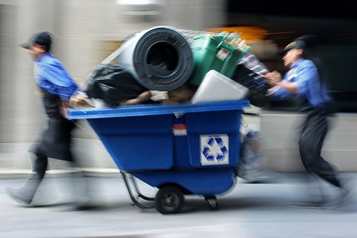 Two waste collection workers, dressed in blue uniforms and black caps, are pushing a large blue recycling bin filled with various rubbish items along a city street. The bin is overloaded with waste materials, including black bin liners, a green plastic container, and a white item that appears to be a household appliance or part of one. The setting suggests an urban environment with a blurred background indicating movement and activity, typical of busy town or city centers such as High Wycombe. The image captures the manual effort involved in rubbish removal as part of commercial waste management services, with the bin positioned on a paved sidewalk or street. The waste appears to be a mixed collection of refuse, and the workers are focused on transporting it for disposal or recycling, consistent with the services offered by Commercial Waste High Wycombe in the local area. The lighting is natural, indicating daytime, with clear visibility of the scene's details and textures, emphasizing the practical aspect of rubbish collection operations in the town.