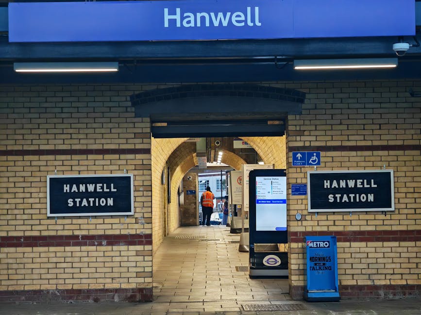 The image depicts the entrance to Hanwell Station, facing a brick building with a large blue sign reading 'Hanwell' at the top. Below the sign, two black and white rectangular signs display 'Hanwell S